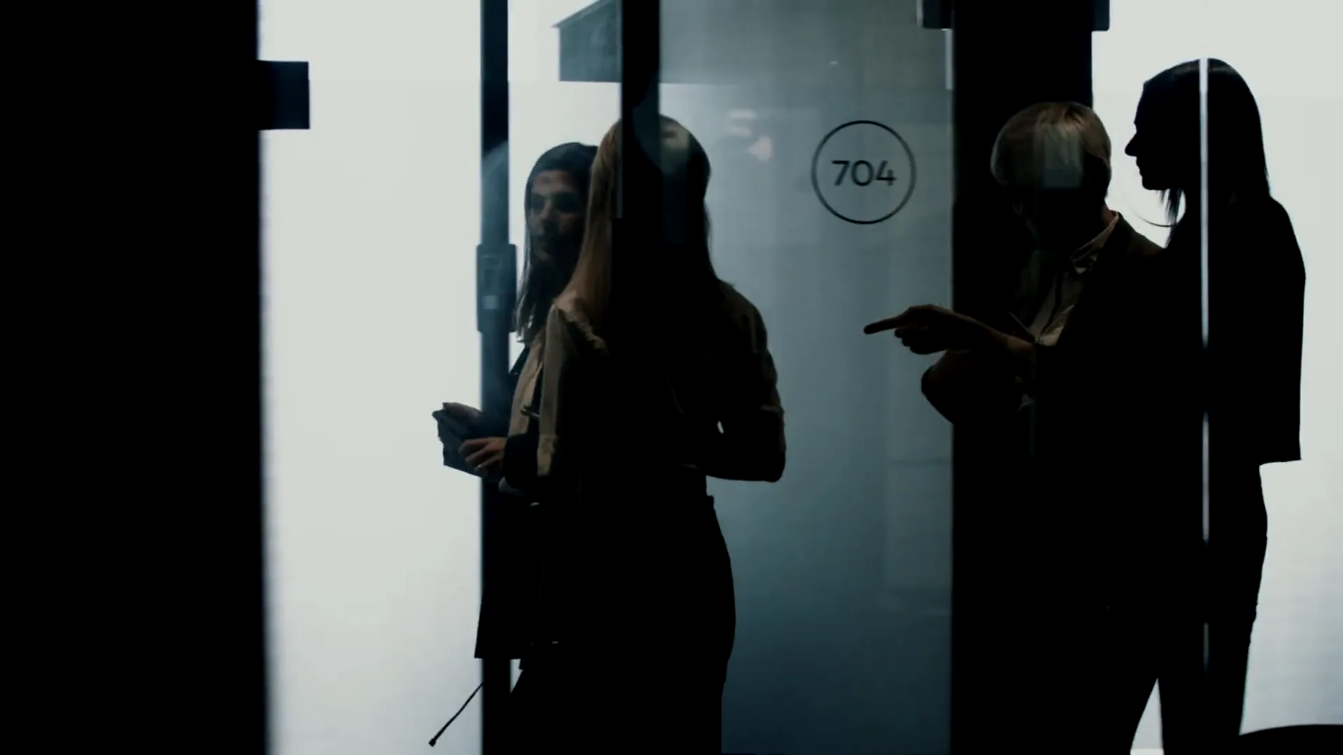 Four colleagues in silhouette at a glass meeting-room door, one pointing as she speaks.