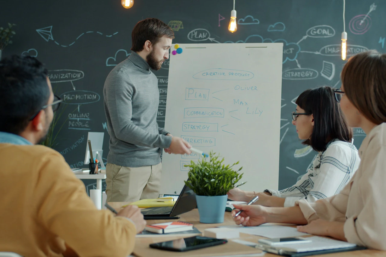 Colleagues at a whiteboard in a meeting room, one standing to explain, two seated with notes.