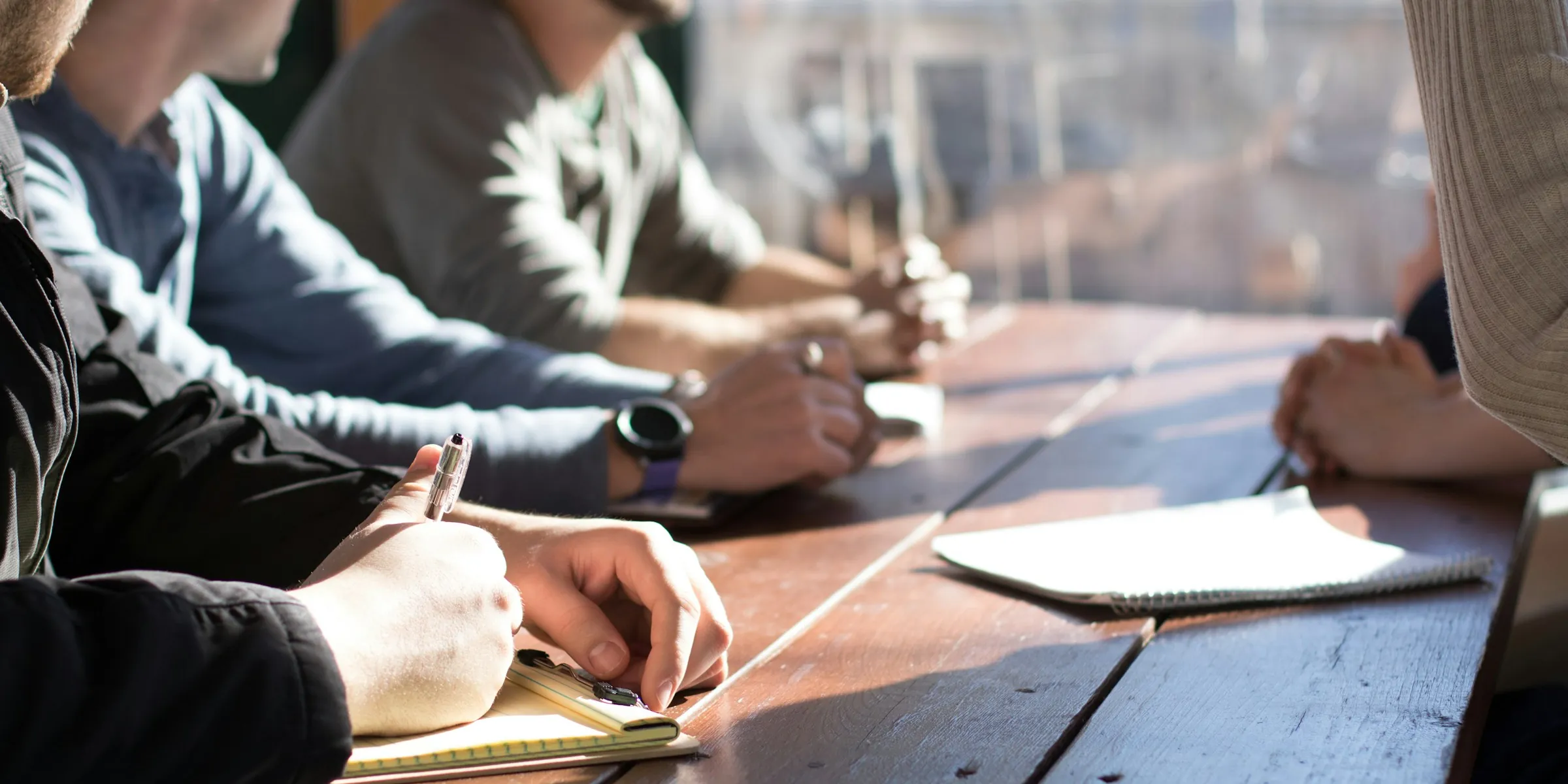 People seated along a long wooden table, notebooks open, one taking notes.