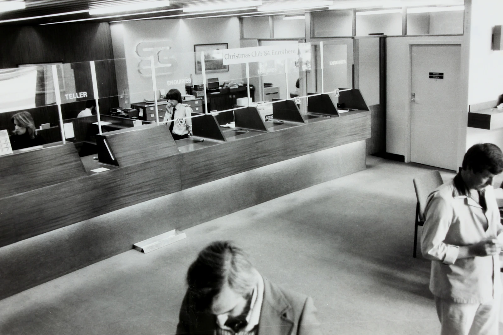 A black-and-white view of a teller counter with glass partitions and staff behind it.