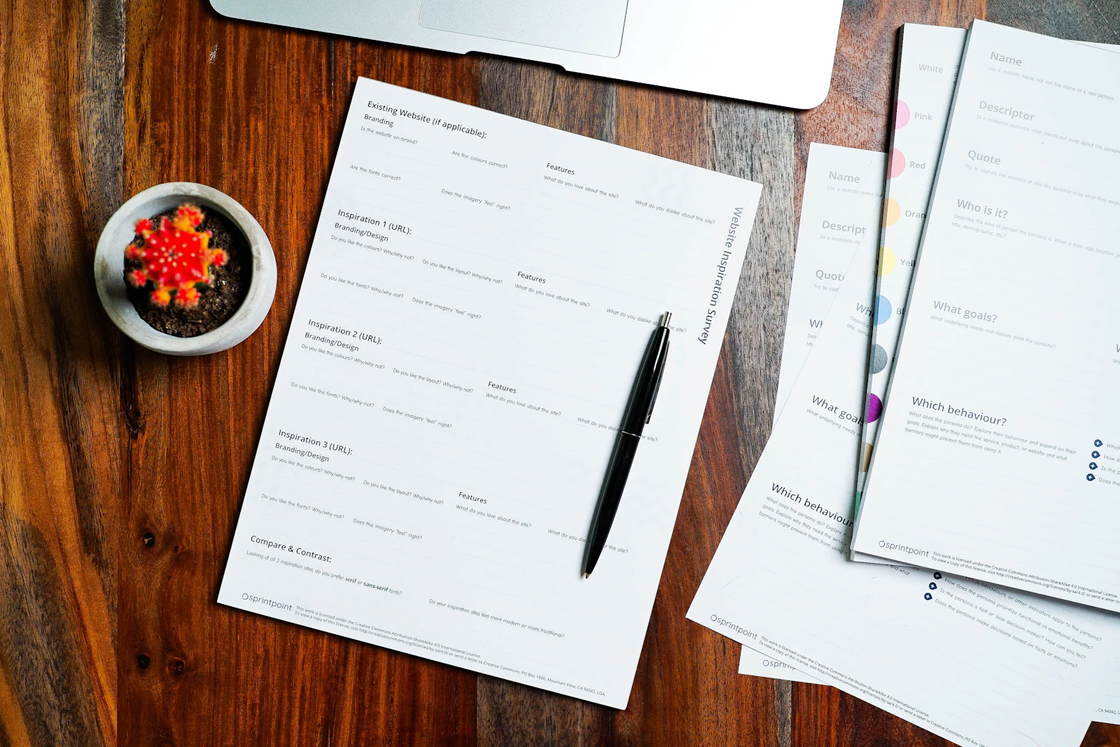 Printed forms, a pen and a laptop on a wooden desk, seen from above.
