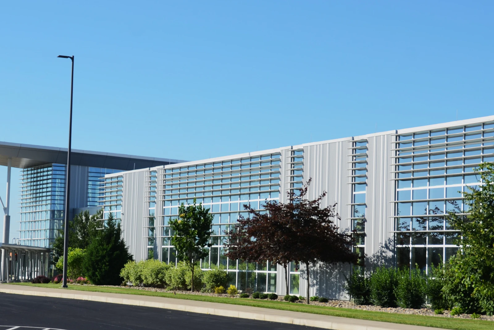 A long glass and metal office building seen from the road, trees and a lamp post in front.