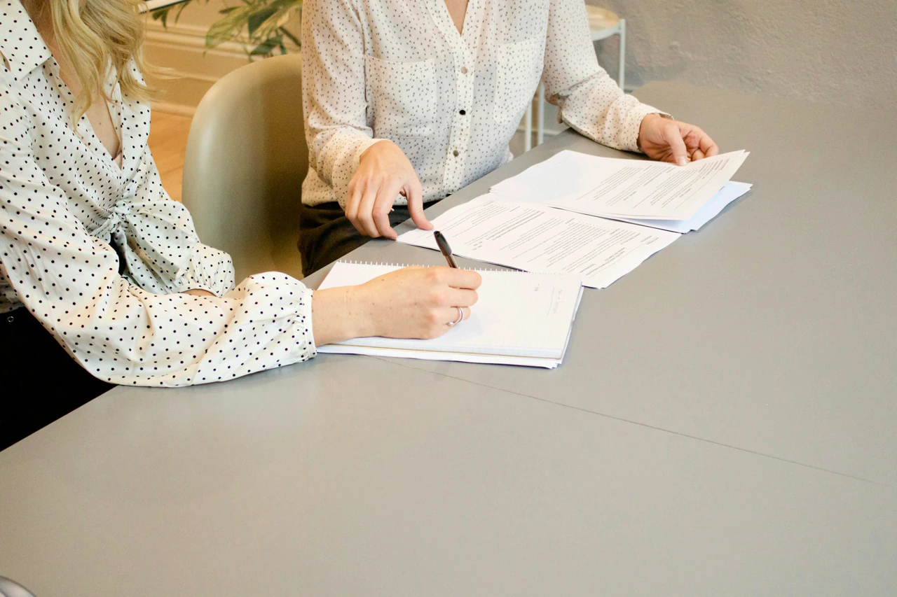 Two professionals across a conference table, documents between them, faces turned to the papers.